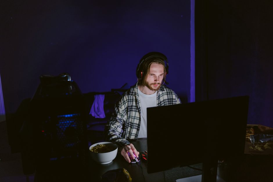 A focused man playing video games on a computer at night with a headset.