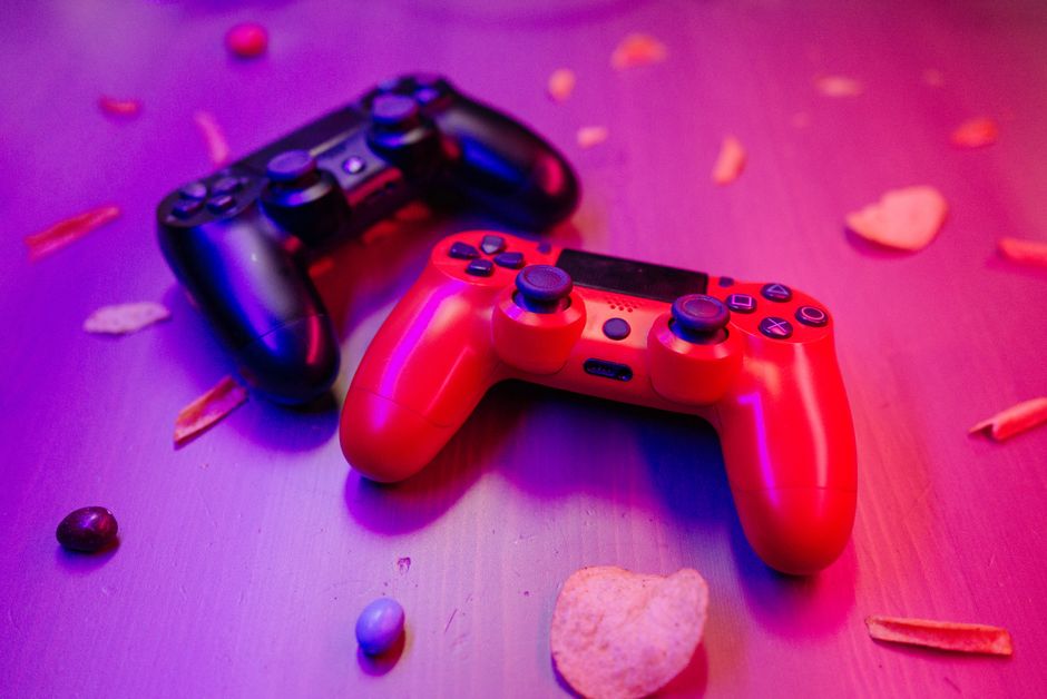 Close-up of red and black game controllers on a table with snacks around, lit by ambient colored light.