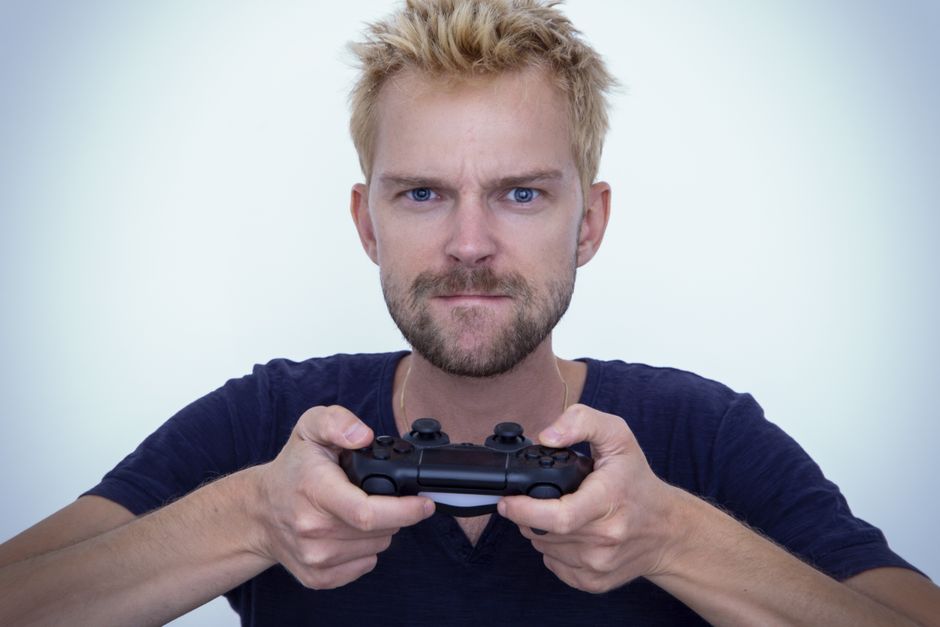 Man with short blonde hair fiercely focused while gaming with a controller against white background.