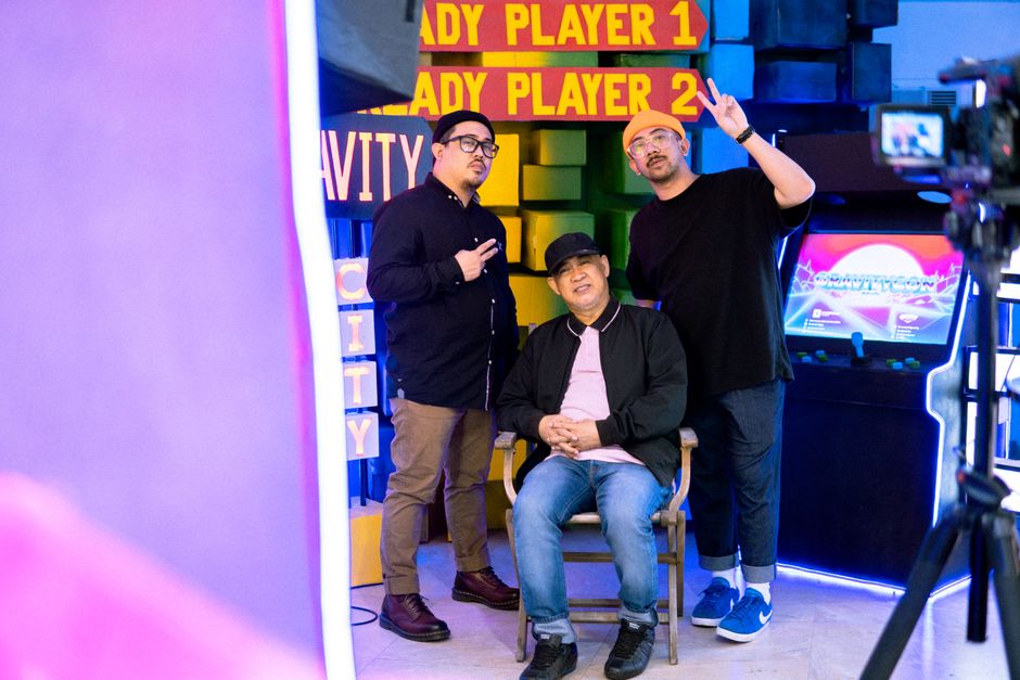 Three fashionable men pose in a vibrant gaming-themed arcade setting filled with retro signs and colorful decor.