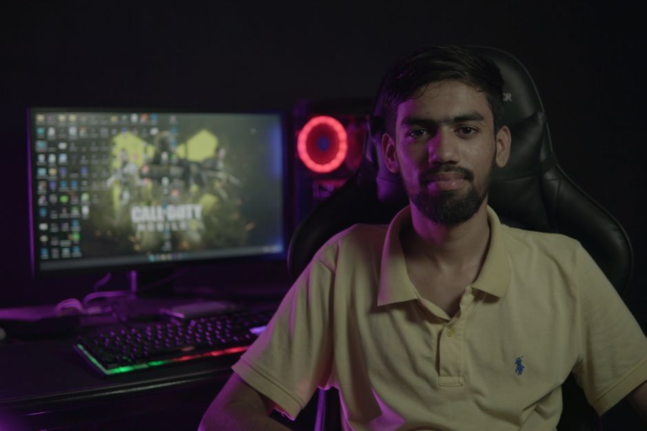A bearded man sitting at a computer setup with a gaming PC and chair, creating a moody ambiance.