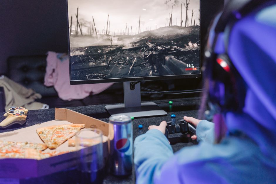 Teen playing video game with headset and drinks, indulging in pizza during gaming session.
